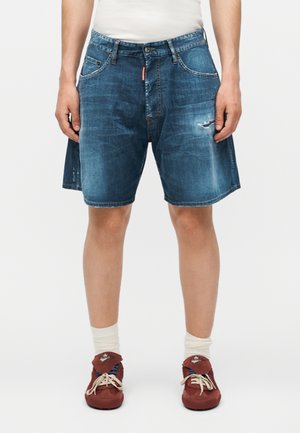 Mand iført blå slidte denimshorts, hvide sokker, røde afslappede sko, stående mod en ensfarvet baggrund.