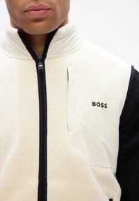 Γούνινο γιλέκο σε κρεμ και μαύρο, με ψηλό γιακά, εμπρός φερμουάρ και μαύρο λογότυπο στο στήθος. Λείος υφασμάτινος χαρακτήρας με αντίθετα μανίκια.