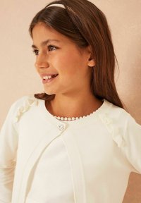 Fehér kardigán fodros részletekkel és drágakő gombos záródással, gyöngy nyaklánccal párosítva, egy hosszú barna hajú lányon.