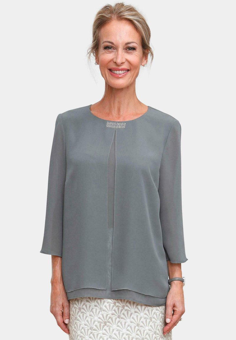 Graue Bluse mit drei Viertelärmeln, Rundhalsausschnitt, vorne mit Falte und Nietenakzenten nahe des Halsausschnitts. Aus glattem, leichtem Material.