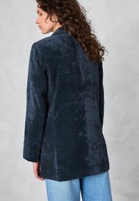 Kvinna med lockigt hår, klädd i en mörkbl�å kordjacka och ljusblå jeans, vänd åt vänster mot en enkel ljus bakgrund.
