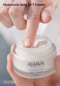 Hyaluronsyra 24-7 kräm i en klar, rund burk med ett vitt lock. Ett finger dyker ner i den krämiga, lätta konsistensen. Märkesnamnet är synligt.