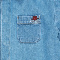Lyseblå denimskjorte med en brystlomme som har rød og hvit brodert tekst og et lite rødt hatt-desegn.