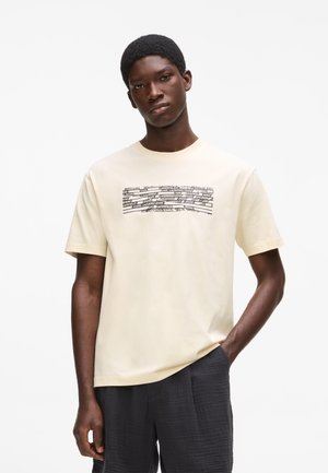 Fiatal sötét bőrű férfi, aki krémszínű pólót visel fekete szöveges grafikával és fekete texturált rövidnadrágot, egyszínű háttér előtt áll.