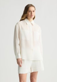 Camicia di lino bianca con maniche lunghe, colletto e due tasche sul petto, abbinata a pantaloni corti in lino coordinati. Texture leggera e traspirante.