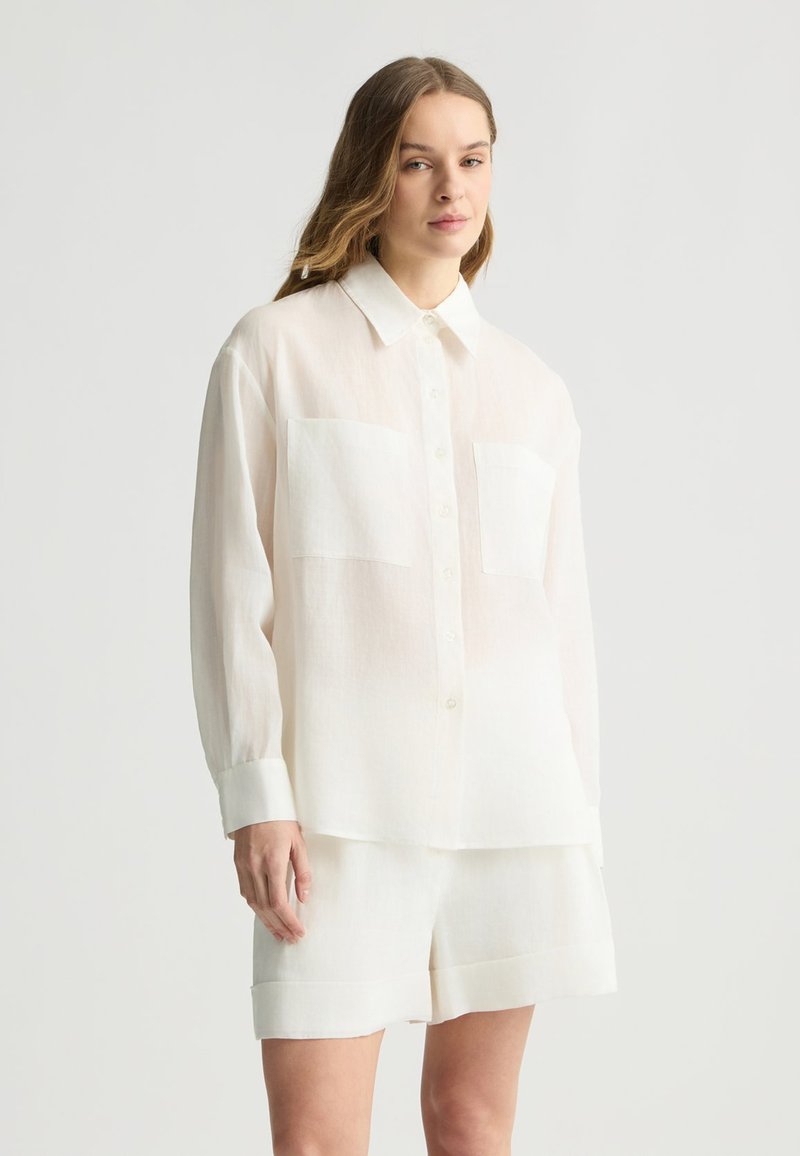 Camicia di lino bianca con maniche lunghe, colletto e due tasche sul petto, abbinata a pantaloni corti in lino coordinati. Texture leggera e traspirante.