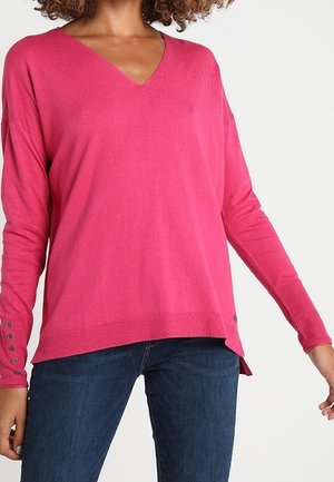 Femme portant un pull à col en V rose vif à manches longues avec des détails de boutons aux poignets et un jean bleu foncé sur un fond uni.
