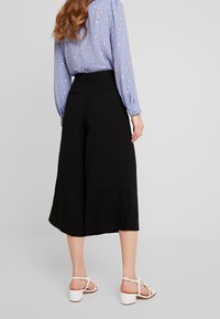 Kvinna klädd i svarta vida culottbyxor, ljusblå blus med prickigt mönster och vita blockhälsandaler, stående med ryggen mot kameran.