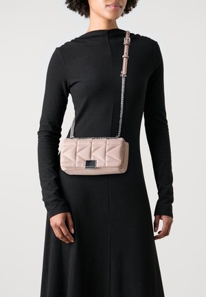 Borsa a tracolla in suede rosa con catena argentata e forma rettangolare. Presenta un piccolo logo sulla parte anteriore. Indossata a tracolla sopra un vestito nero.