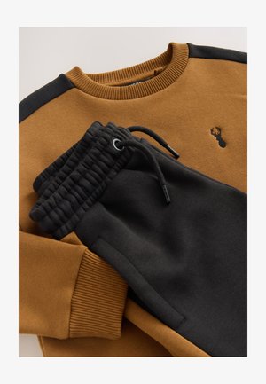 Sweat-shirt marron avec accents noirs aux épaules et petit logo brodé, associé à un pantalon de jogging noir avec taille à cordon et poignets côtelés.