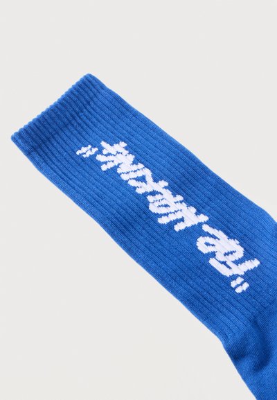 Chaussette bleue côtelée avec texte cursif blanc tricoté le long du côté indiquant "Restez heureux" sur un fond clair uni.