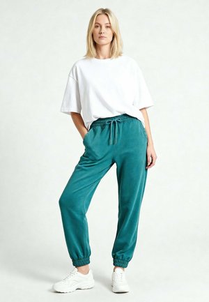 Femme blonde debout, les mains dans les poches de son pantalon de jogging bleu sarcelle, portant un t-shirt blanc ample et des baskets blanches sur un fond clair uni.