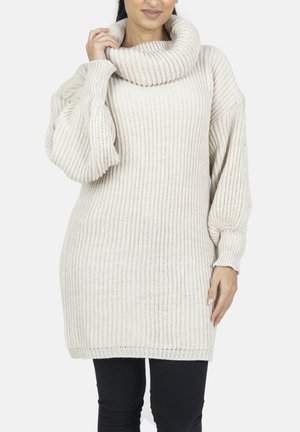 Grober Strickpullover in hellem Beige mit Kropfkragen, Raglanärmeln und gerippter Struktur; lockere Passform, kombiniert mit schwarzen Leggings.