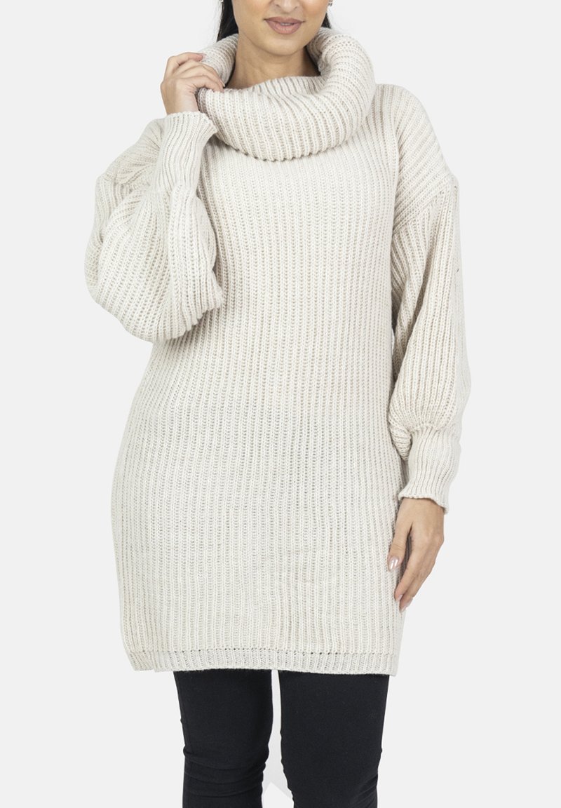 Chunky striktrøje i lys beige med rulledragt, hængende skuldre og ribbet tekstur; løs pasform, kombineret med sorte leggings.