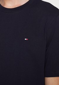 Granatowa bawełniana koszulka z okrągłym dekoltem i krótkimi rękawami, z małym haftowanym logo w kolorach czerwonym, białym i niebieskim na piersi.