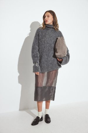 Grauer, flauschiger Pullover mit hohem Kragen, über einem halbtransparenten Rock in gedämpften Farbtönen getragen, kombiniert mit schwarzen Loafers und einer taupefarbenen Tasche.