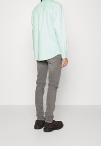 Camisa de manga larga verde menta con detalles de botones, combinada con jeans grises ajustados y zapatos de ante marrón oscuro, vista desde atrás.