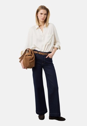 Donna che indossa una camicia bianca a maniche lunghe, jeans larghi scuri, cintura e scarpe marroni, che tiene una grande borsa marrone in suede con la mano in tasca.