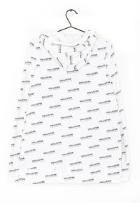 Vit hoodie med ett heltryckt mönster i svart med texten "Hollister" och "California". Mjuk tyg, långa ärmar och en huva med dragsko.