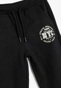 Pantalones de chándal negros hechos de tela suave, con una cinturilla elástica y cordones, y un gráfico circular blanco etiquetado como "NYC."