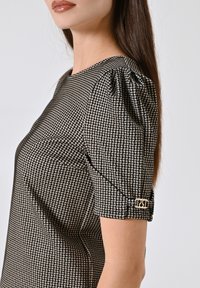 Top a fantasia houndstooth bianco e nero con maniche a sbuffo e un bottone dorato con le lettere "RST." Texture liscia.