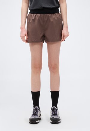 Persoană care poartă pantaloni scurți maro, șosete negre și pantofi sport gri, stând pe un fundal alb simplu.