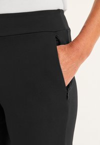 Zwarte slim-fit broek met een ritszak, gladde stof en een strakke uitstraling. Zichtbare hand gestoken in de zak.