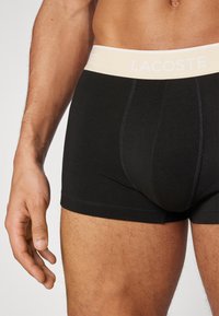 Melni bokseri šorti, izgatavoti no kokvilnas, ar pelēku elastīgu jostasvietu, uz kuras ir redzams "LACOSTE" logotips baltā krāsā. Uz auduma redzamas šuvju detaļas.