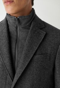 Blazer grigio a spina di pesce con un maglione a collo alto e zip sotto. Tessuto strutturato, vestibilità sartoriale e design minimalista.