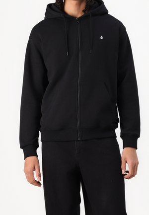Personne portant un sweat à capuche noir zippé avec un petit logo blanc sur la poitrine gauche et un pantalon noir sur un fond blanc uni.
