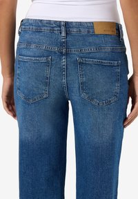 Jeans din denim albastru cu un croi drept, având două buzunare pe spate și o etichetă maro pe talie. Material texturat cu un finisaj decolorat.