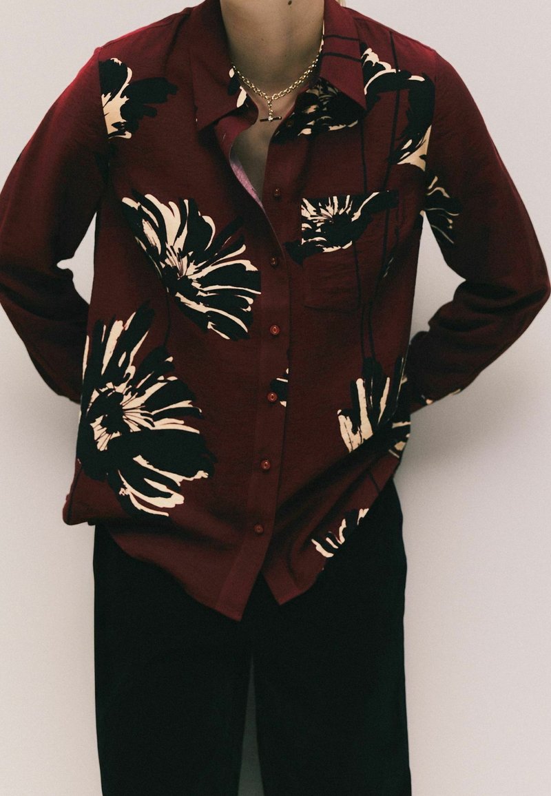 Personne portant une chemise bordeaux boutonnée avec de grandes impressions florales noires et blanches et un pantalon noir, debout les mains derrière le dos.