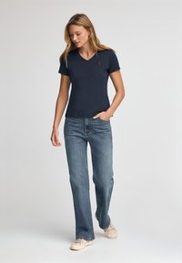 Marinblå V-ringad t-shirt i mjukt material, ihopkopplad med ljusblå utsvängda jeans och beige sneakers. Röd logotypdetalj på skjortan.