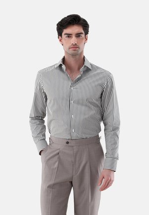 Mann mit dunklen Haaren, der ein schwarz-weiß vertikal gestreiftes Hemd trägt, das in hochtaillierte hellbraune, plissierte Hosen gesteckt ist, steht mit einer Hand in der Tasche.