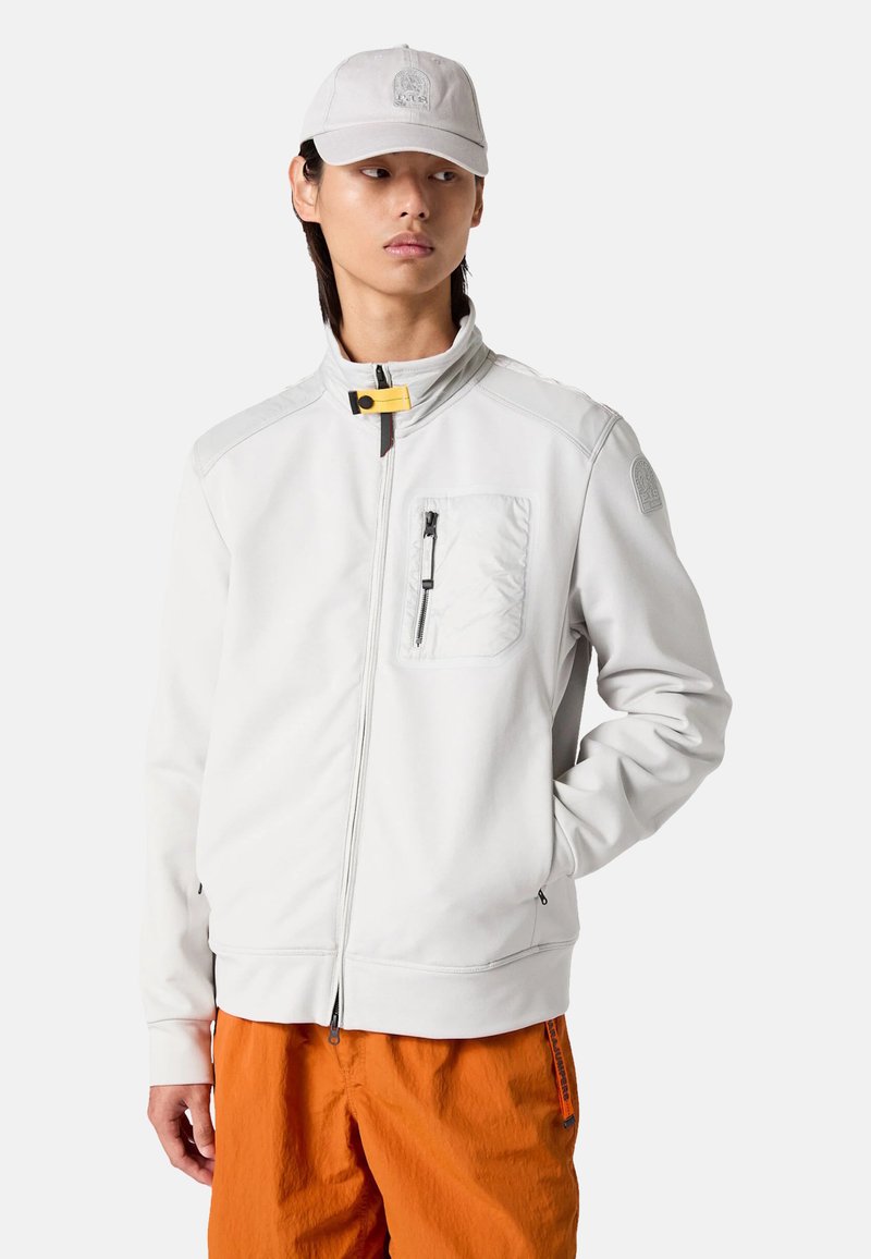 Weiße Zip-Jacke mit hohem Kragen, einer Brusttasche, schwarzem Reißverschluss und einem Logo-Patch auf der Schulter, kombiniert mit orangefarbenen Hosen.