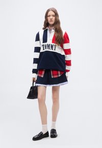 Γυναίκα που φοράει πουλόβερ Tommy με ρίγες σε μπλε σκούρο, λευκό και κόκκινο, με πουκάμισο καρό από μέσα, μπλε σκουρό χρώμα φούστα, λευκές κάλτσες, μαύρα μοκασίνια, κρατώντας μια μαύρη τσάντα χειρός.