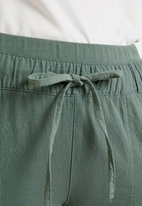 Pantalones de tiro alto en un verde claro con cintura elástica y un acabado texturizado, con un lazo en la parte frontal. La banda de la cintura cuenta con detalles acanalados para mayor comodidad.