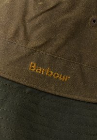 Κοντινή λήψη του καφέ κερωμένου υφάσματος με το ραμμένο πορτοκαλί λογότυπο "Barbour" και σκοτεινό πράσινο κουβερτίνα κάτω.