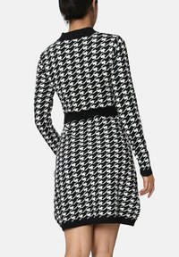 Femme portant une robe à manches longues en motif pied-de-poule noir et blanc avec une taille et un col noirs, vue de dos.