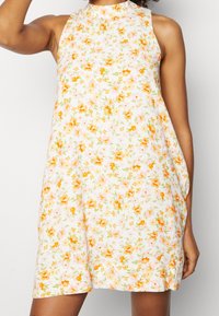 Blommig klänning med hög halsringning, som har en vit bas prydd med små orange och rosa blommor, samt en lös A-linjeform.