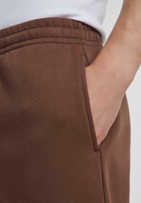 Brune sweatpants har en glat tekstur, elastisk talje, og en sidelomme. Stoffet synes blødt og har et afslappet design.