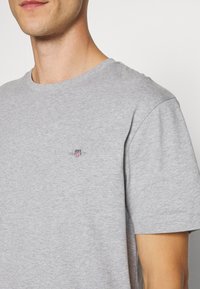 Siva bombažna majica s krožnim izrezom in kratkimi rokavi. Na levi strani prsi ima majhen vezen logotip. Gladek tekstura.