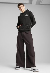 Felpa nera con tasca frontale e logo bianco, abbinata a pantaloni larghi marroni scuri e sneakers bianche con dettagli neri.