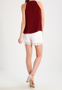 Bordowy bezrękawnik z zaokrąglonym dołem w połączeniu z białymi koronkowymi szortami z falbaniastymi brzegami. Neutralne szpilki dopełniają stylizację.