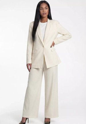 Kobieta stojąca z ręką na biodrze, ubrana w kremowy garnitur w prążki z szerokimi spodniami i czarne buty na wysokim obcasie, na białym tle.