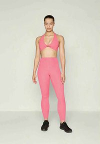 Žena stojící v růžovém sportovním topu a legínách s vysokým pasem, s černými sportovními botami na pozadí v jednoduché světlé barvě.