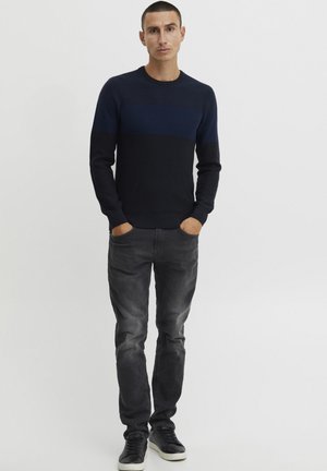 Pull en tricot bleu marine à rayures horizontales ; manches longues, col rond ; associé à un jean gris et des baskets noires.