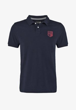 Polo de color azul marino hecho de algodón, con cuello de canalé, tapeta de dos botones y un emblema rojo en el lado izquierdo del pecho.