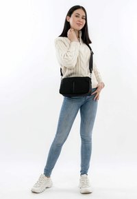 Borsa a tracolla nera in tessuto liscio, con tasca frontale con zip e tracolla regolabile. Il modello indossa un maglione crema e jeans blu.