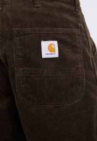 Brune fløjl bukser med lodrette ribber, en baglomme og en hvid label med et orange logo. Struktureret stof med robust design.